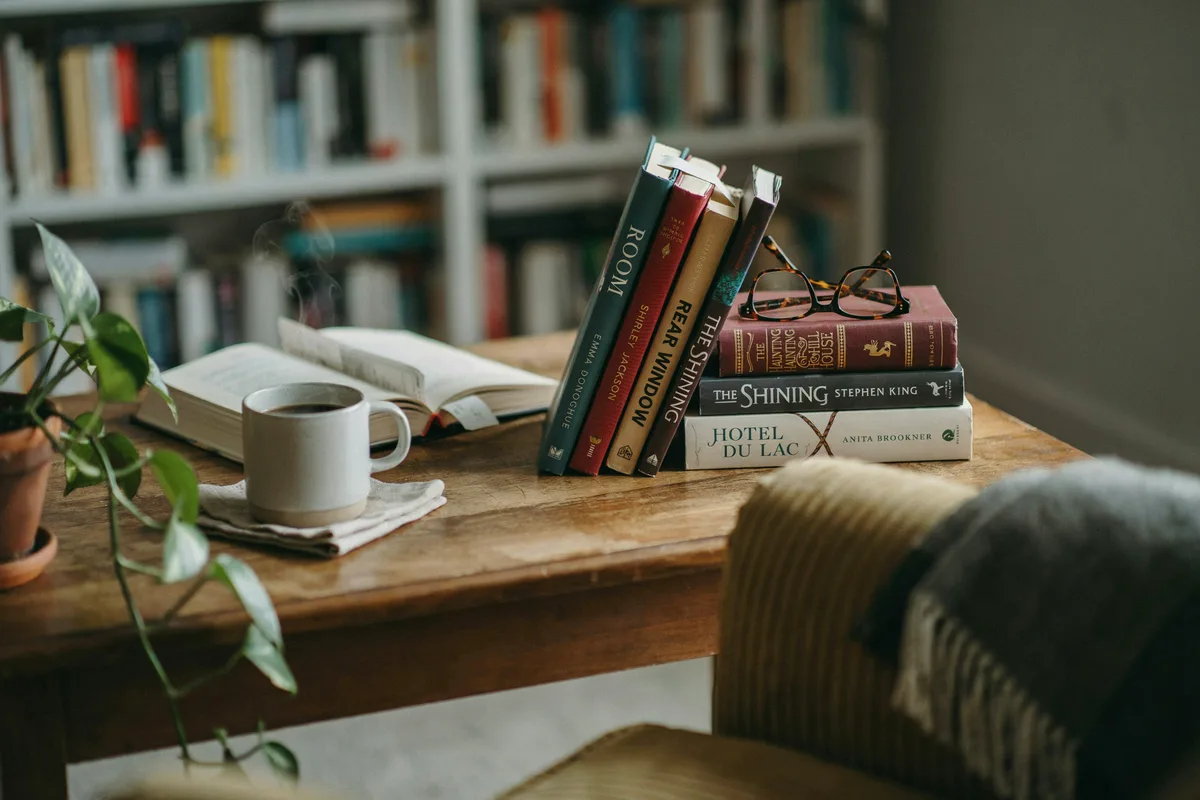 A stack of novels confined to a single setting, including The Haunting of Hill House and Rebecca, on a desk with reading glasses.