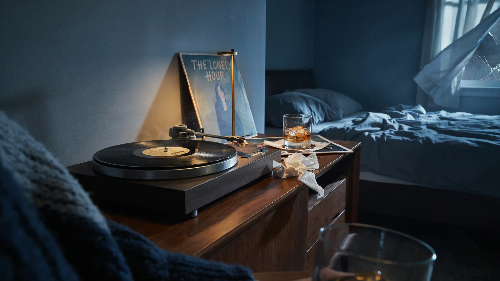 A dim bedroom with a turntable playing a record titled The Lonely Hour beside a whiskey glass and crumpled tissues.
