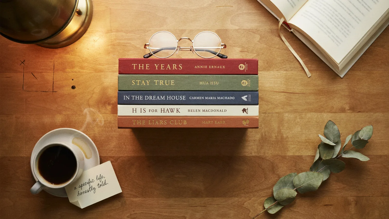 A stack of acclaimed memoirs including The Years and Stay True on a desk with glasses, coffee and a note reading a specific life honestly told.