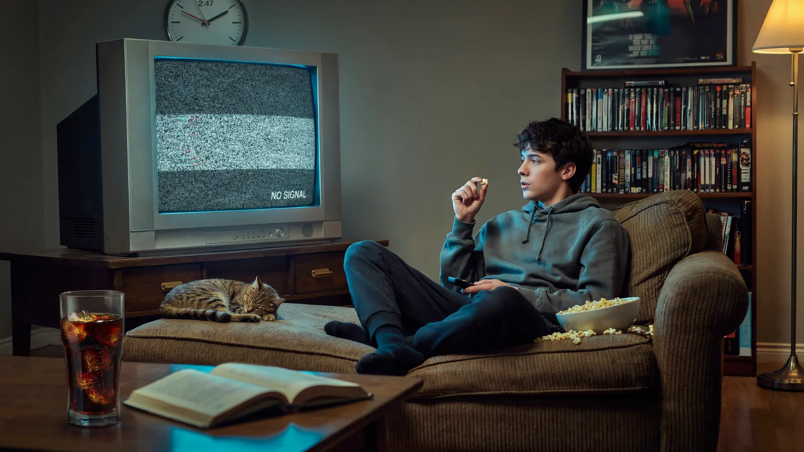 A young man watching static on an old television labeled No Signal from his couch, surrounded by a DVD collection and a sleeping cat.