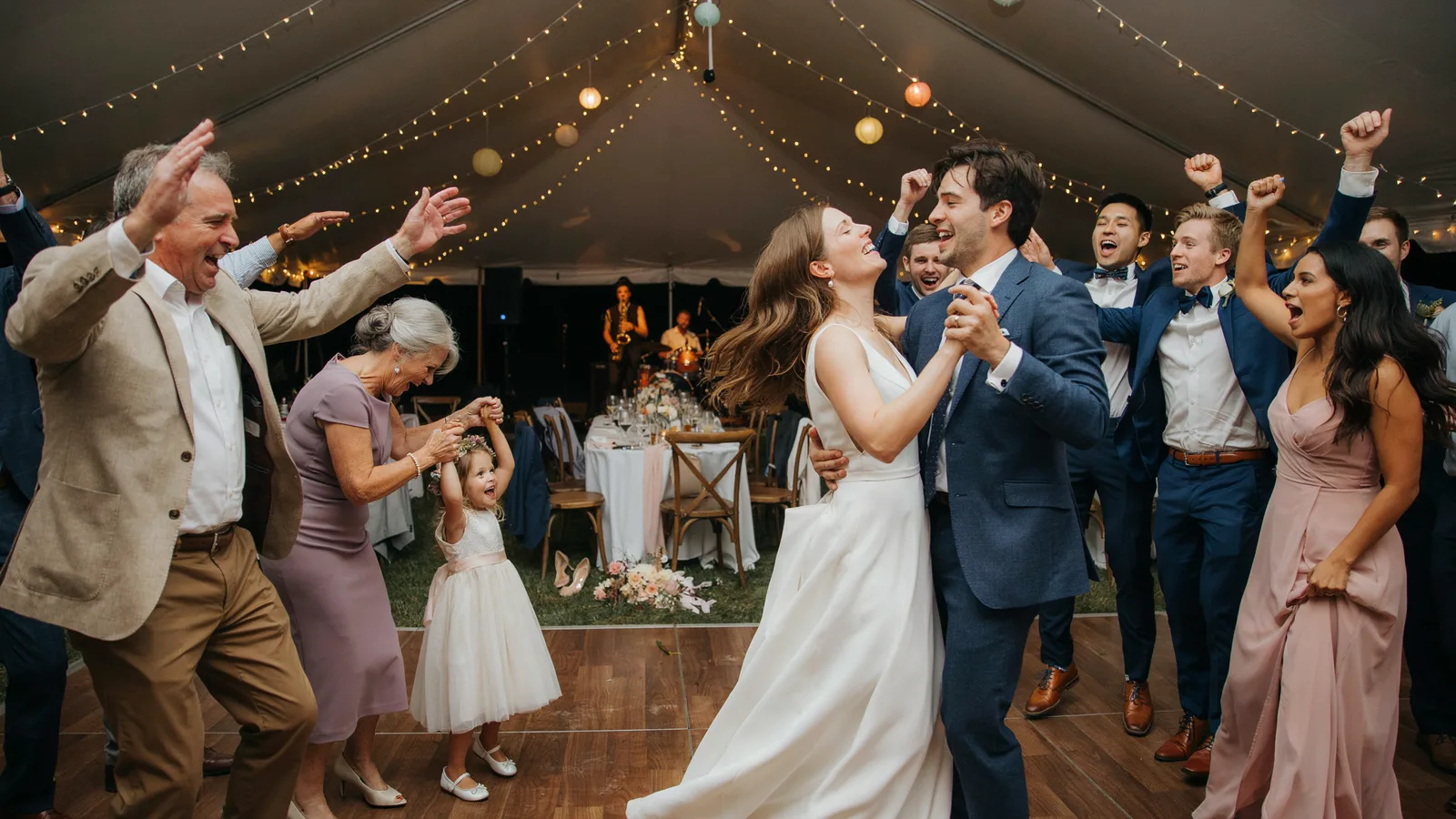 A wedding reception dance floor with the bride, groom and guests cheering under string lights.